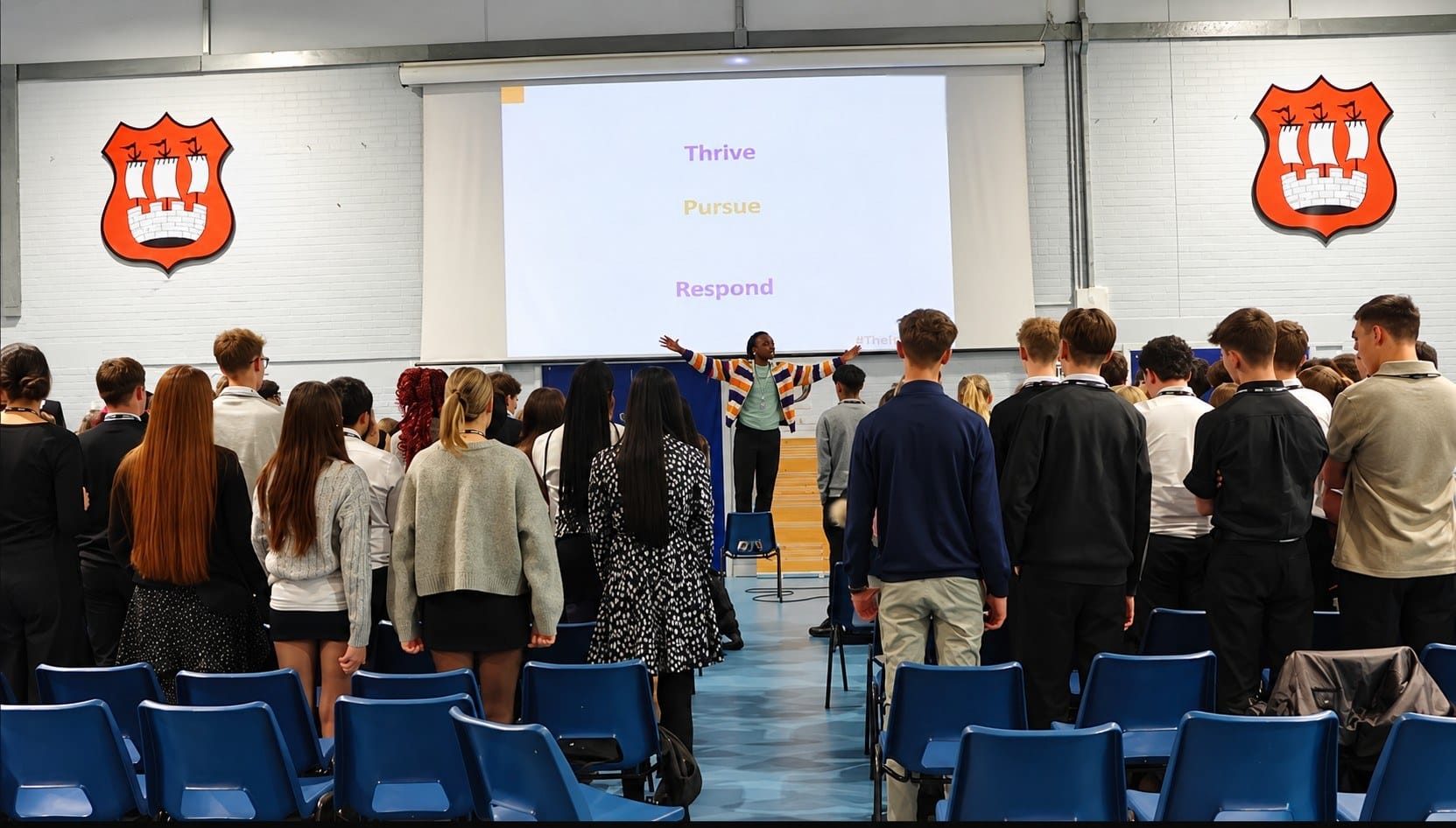 Mike O with both arms wide in a school hall while students stand and listen. The screen behind him reads Thrive, Pursue, Respond.