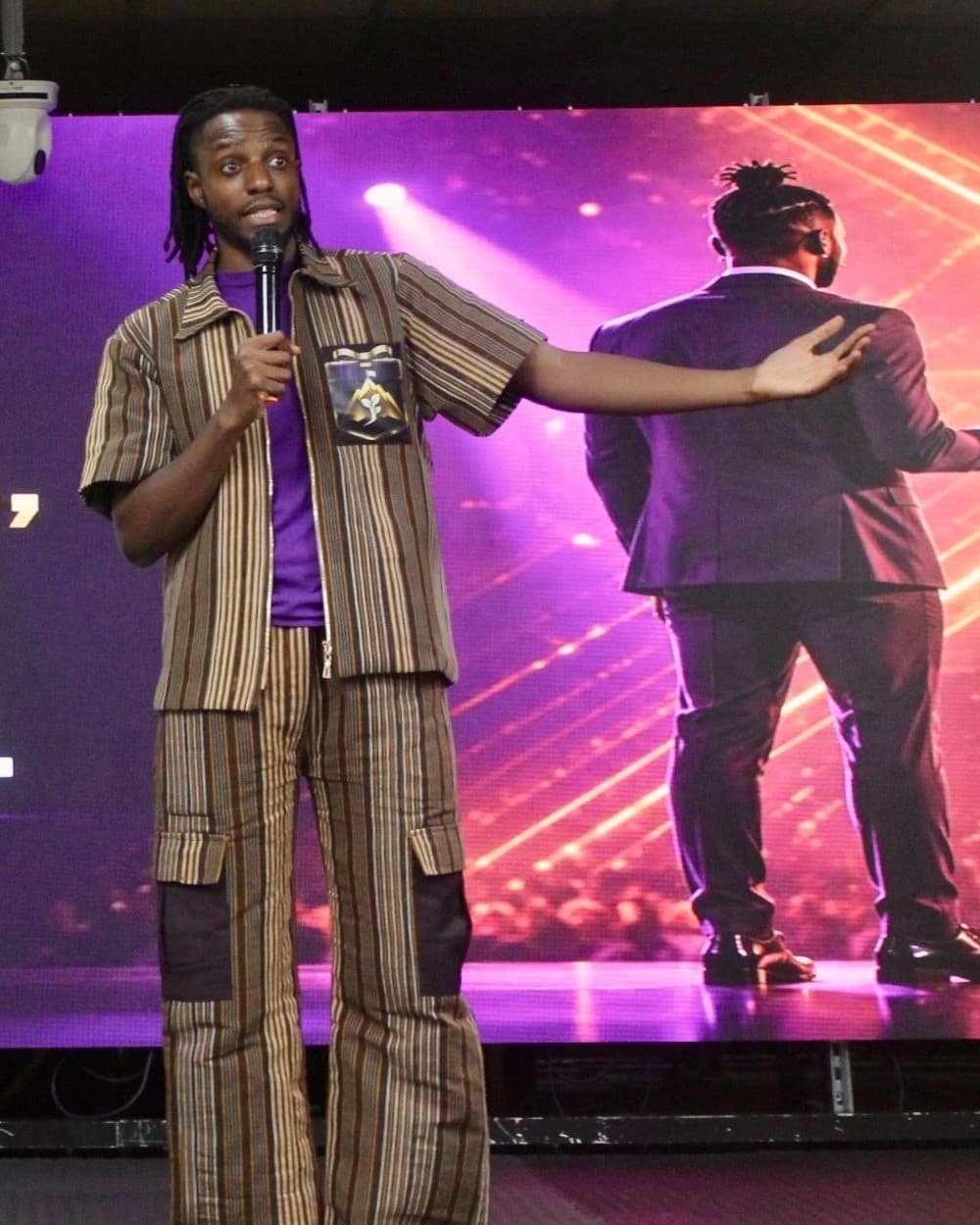 Mike O on stage at an opening keynote, microphone in hand and one arm held out, in front of a large purple screen showing the line How to navigate well, not just perform well.