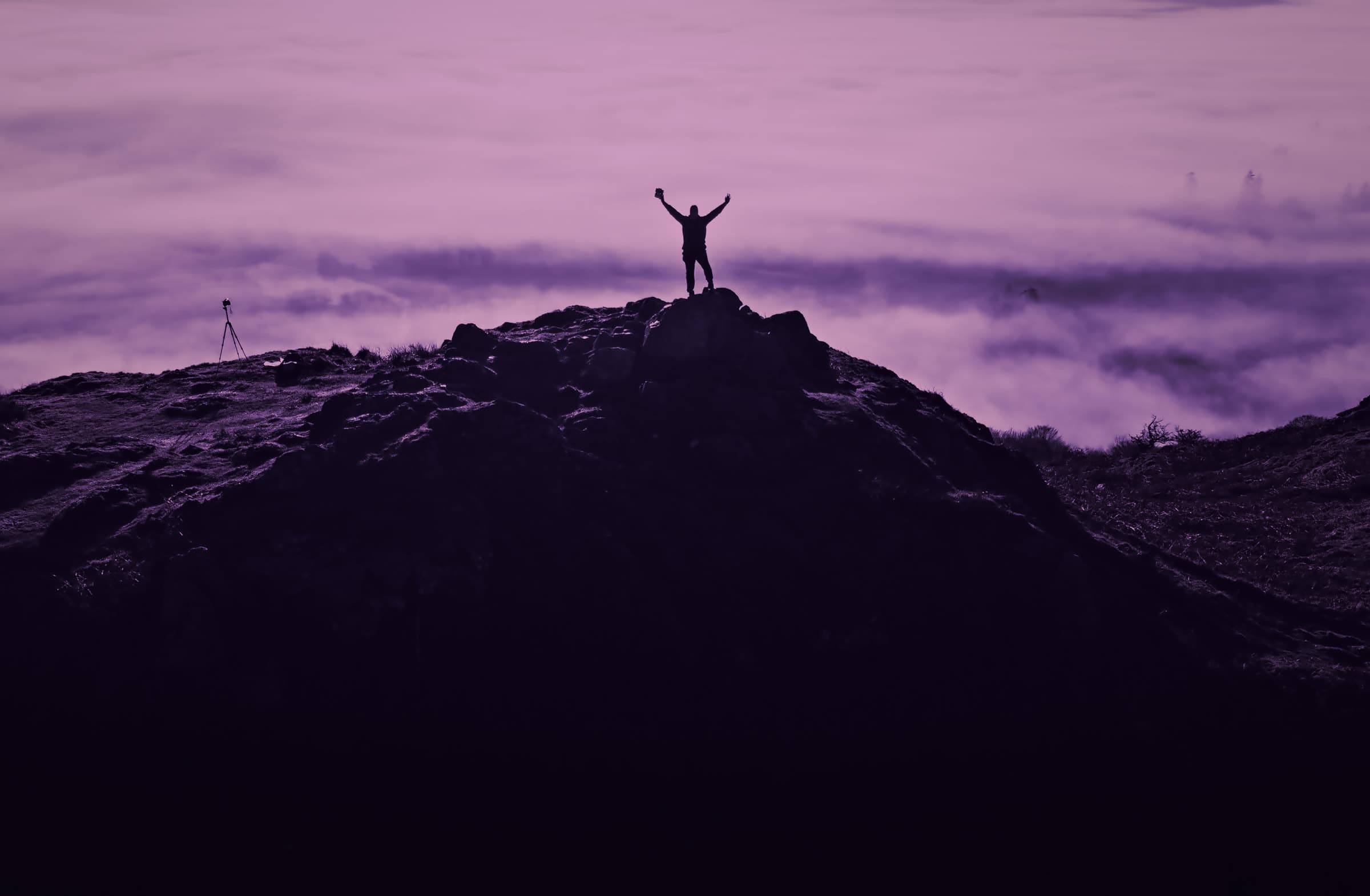 A figure with arms raised on a summit above the clouds at dusk.