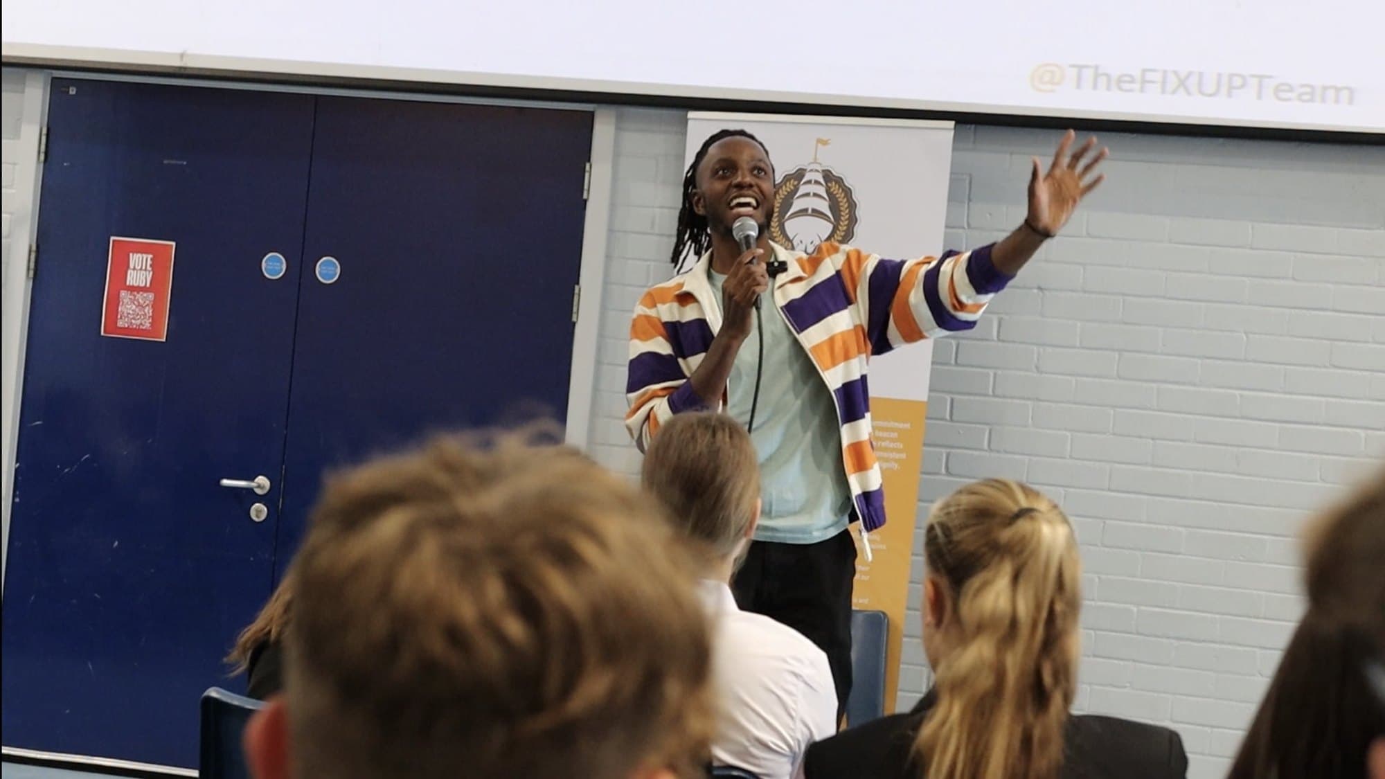 Mike O in a striped jacket speaking into a microphone with one hand raised, in a school hall, with pupils seen from behind in the foreground.